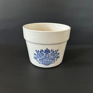 May include: A small, white ceramic planter with a blue floral design. The design features a basket of flowers with leaves and stems.