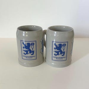 May include: Two light gray ceramic beer mugs featuring a blue rectangular logo with a lion graphic and the text LOWENBRAU MUNICH. Each mug has a sturdy handle on the side and a smooth, cylindrical body with a slightly flared rim and base design.