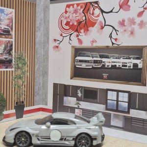 Può includere: Un'auto giocattolo argentata con ruote nere e un cerchio bianco sul lato. Lo sfondo presenta un'immagine incorniciata di auto bianche, arte di fiori di ciliegio e una scena di cucina in miniatura.