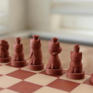 May include: A set of brown, matte-finish chess pieces shaped like cats, each standing on a circular base. The pieces include cat figures wearing crowns or pointed hats, arranged on a checkered board with alternating brown and beige squares.