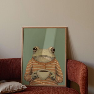 May include: A framed art print featuring a frog wearing an orange and white striped shirt, holding a cup of coffee. The frog is depicted with detailed shading and texture, set against a muted green background. The artwork is framed in a light-colored wooden frame.