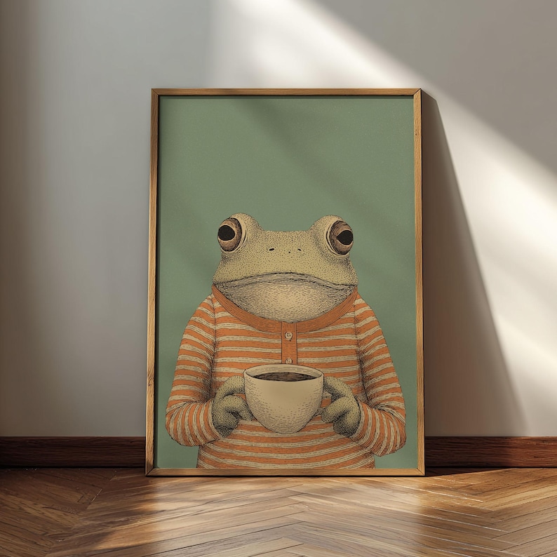 May include: Framed art print featuring a whimsical illustration of a frog wearing an orange and white striped shirt, holding a cup of coffee. The frog is set against a muted green background. The artwork is framed in a natural wood frame.