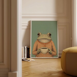 May include: A framed art print featuring a whimsical illustration of a frog holding a coffee cup. The frog wears an orange and white striped sweater against a muted green background. The artwork is displayed in a white frame.