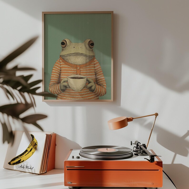 May include: A framed art print featuring a frog wearing an orange striped shirt, holding a cup of coffee. The print hangs above an orange record player with a vinyl record and a banana album cover.