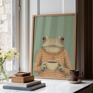 May include: A framed art print featuring a frog wearing an orange and white striped shirt, holding a cup of coffee. The background is a light green with vertical stripes. The artwork is displayed on a white surface near a window.