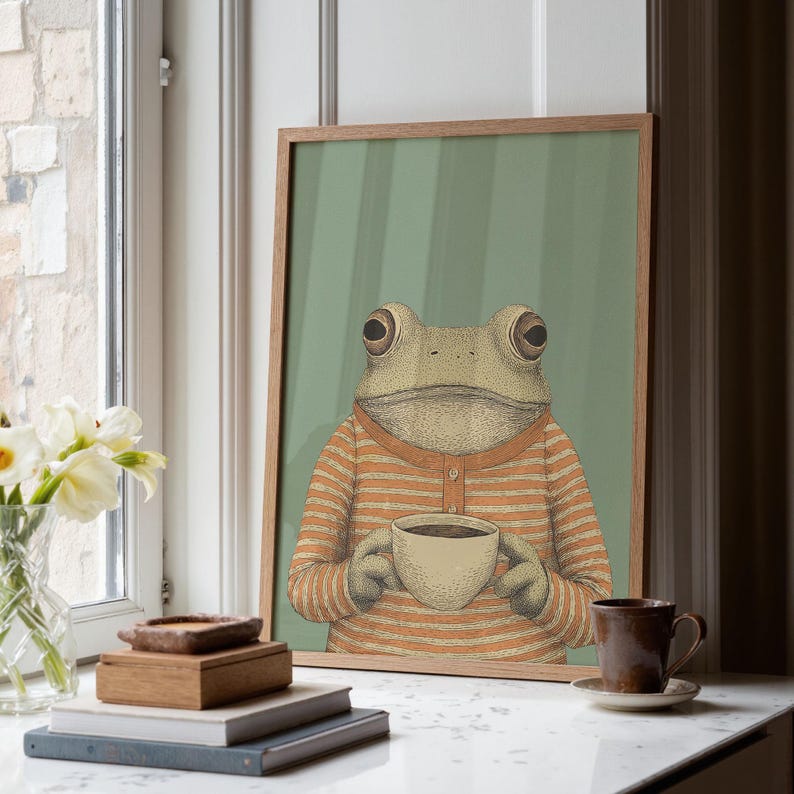 May include: A framed art print featuring a frog wearing an orange and white striped shirt, holding a cup of coffee. The background is a light green with vertical stripes. The artwork is displayed on a white surface near a window.