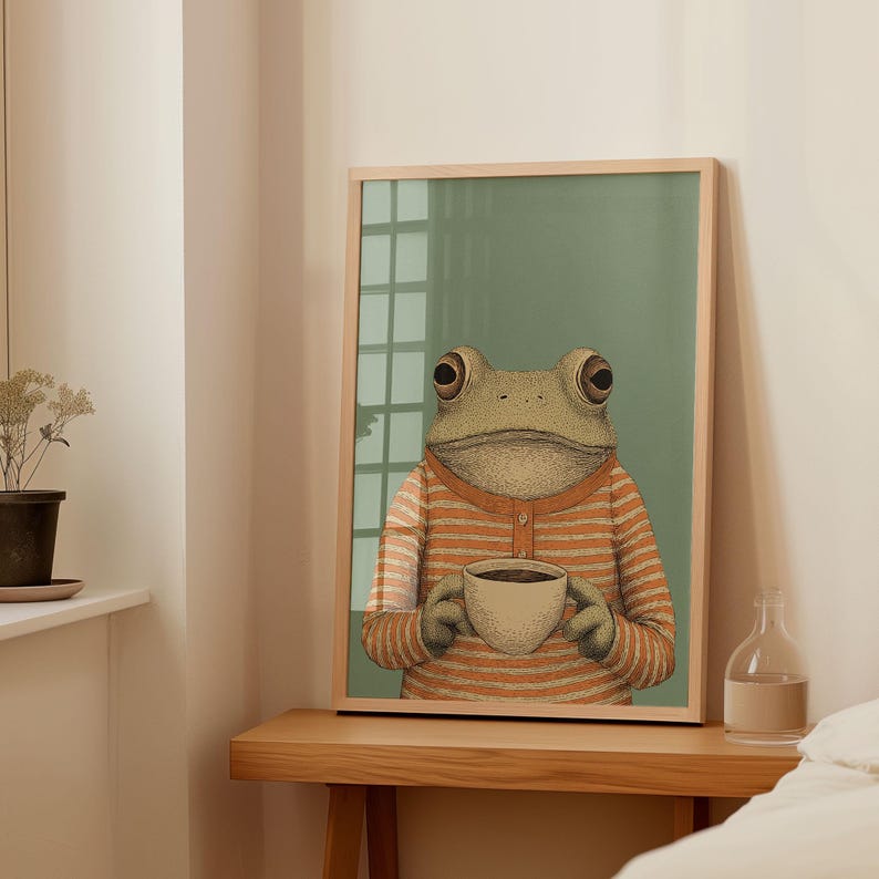 May include: A framed art print featuring a frog wearing an orange and white striped sweater, holding a cup of coffee. The frog has large, expressive eyes and is set against a muted green background. The artwork is displayed on a wooden table.