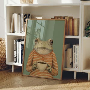 May include: A framed art print featuring a whimsical illustration of a frog wearing an orange striped shirt and holding a coffee cup. The artwork has a light green background and a wooden frame. The print is leaning against a white bookshelf.