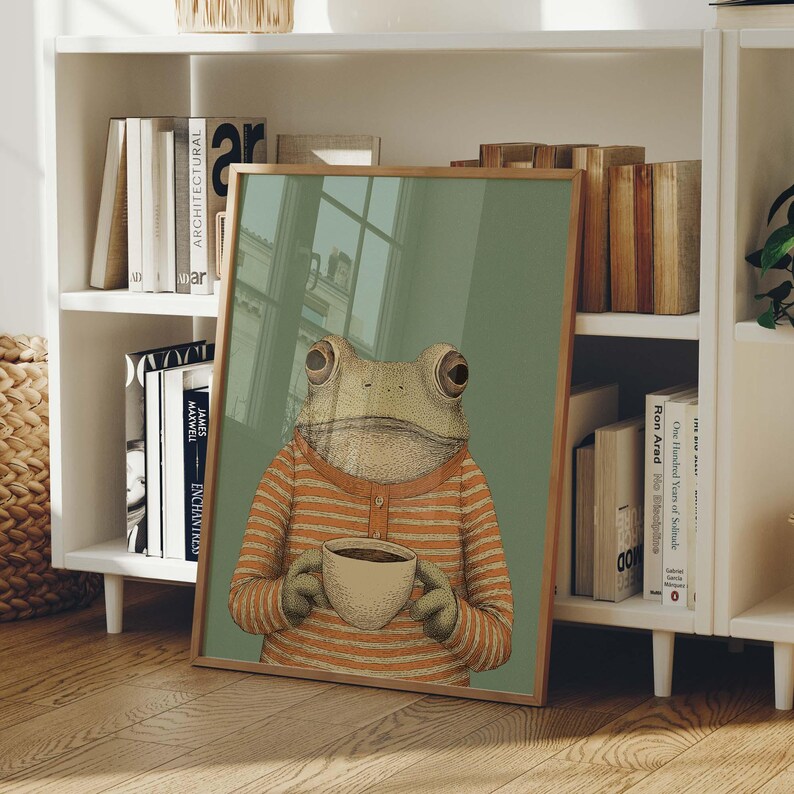 May include: A framed art print featuring a whimsical illustration of a frog wearing an orange striped shirt and holding a coffee cup. The artwork has a light green background and a wooden frame. The print is leaning against a white bookshelf.