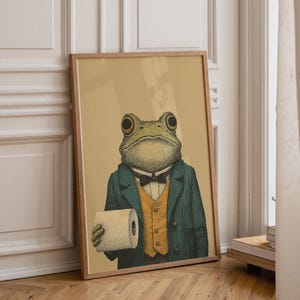 May include: Framed art print featuring a frog dressed in a suit, holding a roll of toilet paper. The frog has large eyes and is wearing a dark green jacket, a yellow vest, and a black bow tie. The background is a light tan color.