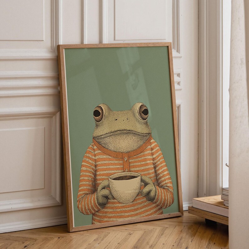 May include: Framed art print featuring a whimsical illustration of a frog wearing an orange and white striped shirt, holding a cup of coffee. The frog is set against a muted green background, creating a cozy and inviting atmosphere. The artwork is in a wooden frame.