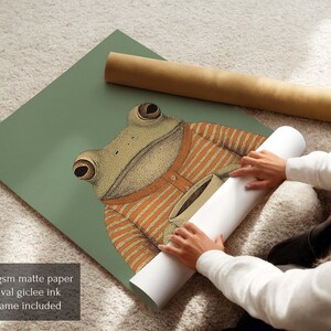 May include: A whimsical illustration of a frog wearing an orange striped sweater, set against a muted green background. The frog is the focal point, with detailed eyes and a friendly expression. A rolled print and a brown tube are also visible.