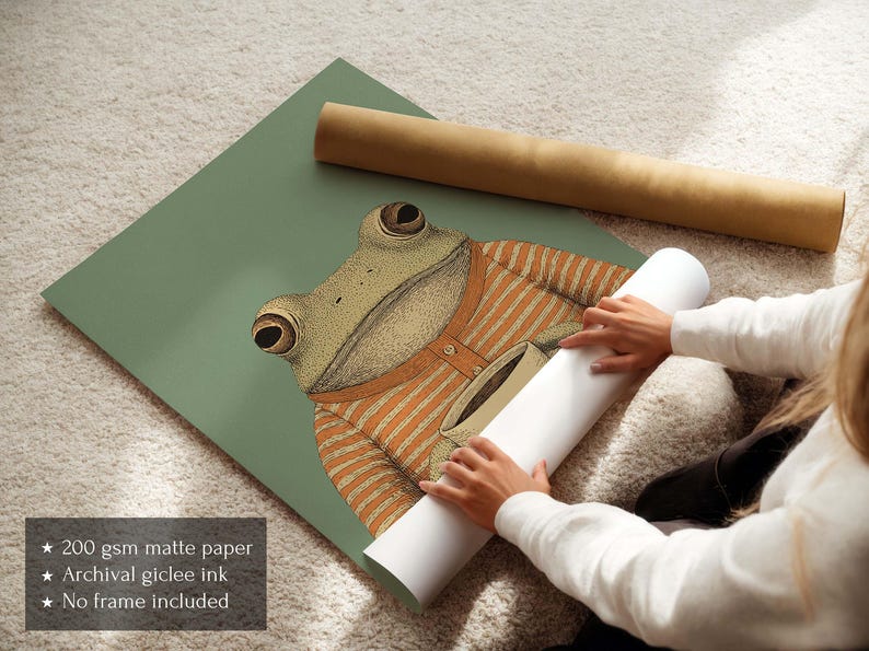 May include: A whimsical illustration of a frog wearing an orange striped sweater, set against a muted green background. The frog is the focal point, with detailed eyes and a friendly expression. A rolled print and a brown tube are also visible.