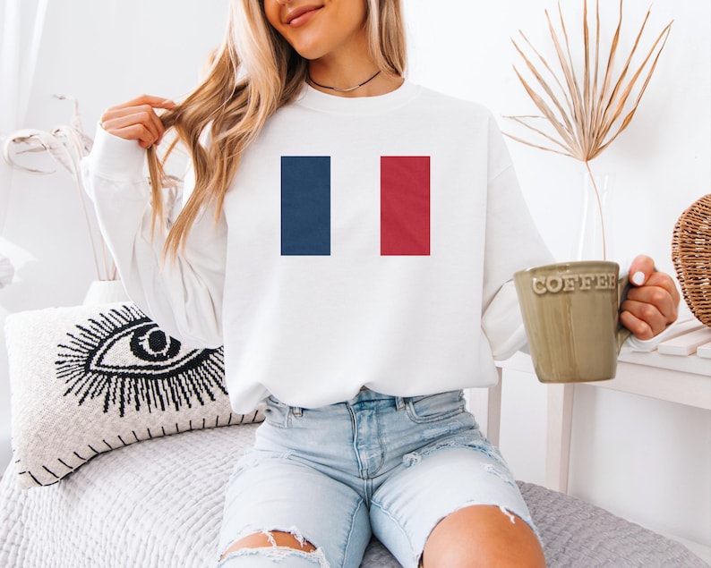 A woman wearing a white sweatshirt with a French flag design is sitting on a bed, holding a coffee mug.