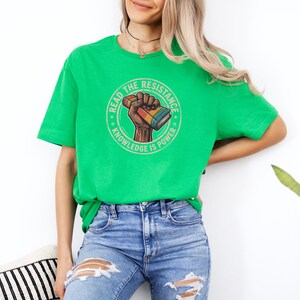 A woman wearing a green t-shirt with a graphic design and jeans stands against a white wall, with a potted plant in the background.