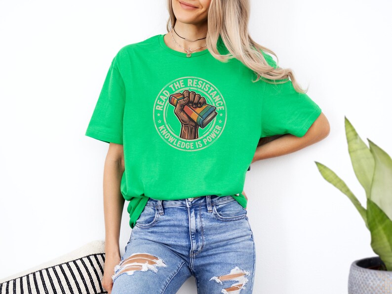 A woman wearing a green t-shirt with a graphic design and jeans stands against a white wall, with a potted plant in the background.