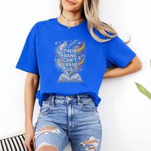 A woman wearing a blue t-shirt with a colorful graphic and distressed jeans stands against a white wall, with a potted plant visible to her right.