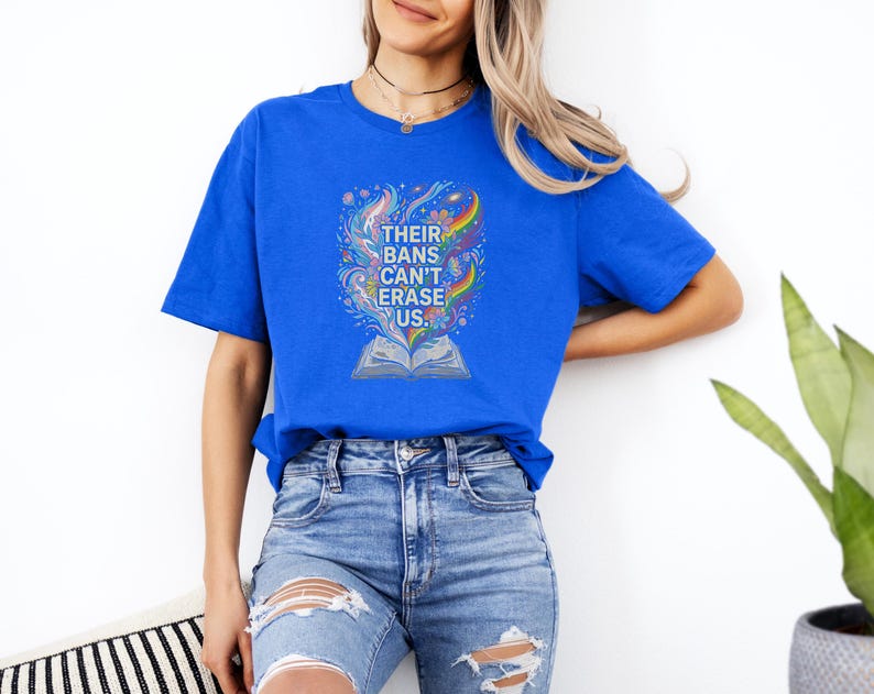 A woman wearing a blue t-shirt with a colorful graphic and distressed jeans stands against a white wall, with a potted plant visible to her right.