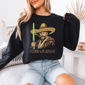 May include: Black long-sleeve sweatshirt featuring a graphic of a mustached man in a sombrero holding a green lightsaber. The text "OBI-JUAN" is printed below the graphic in a gold color.