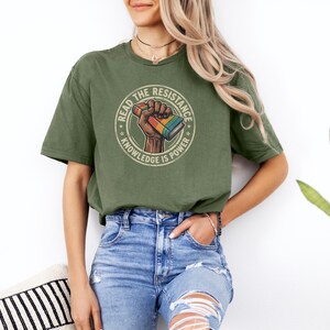 A woman wearing a green t-shirt with a graphic design and jeans stands in front of a white wall, with a potted plant to her right.