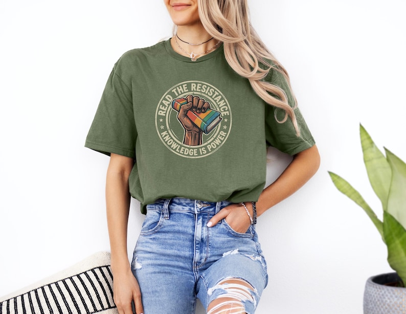 A woman wearing a green t-shirt with a graphic design and jeans stands in front of a white wall, with a potted plant to her right.