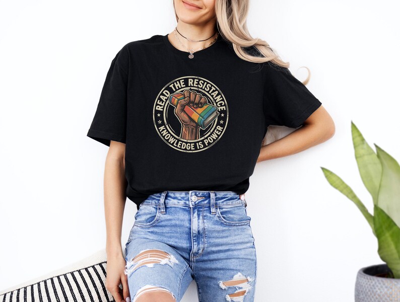 A woman wearing a black t-shirt with a graphic design and jeans stands in front of a white wall, with a potted plant visible to her right.