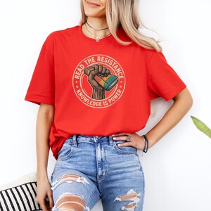 A woman wearing a red t-shirt with a graphic design and jeans stands in front of a white wall, with a potted plant visible to her right.