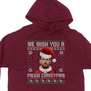 May include: Burgundy hoodie with a festive design. The front features a portrait of a man wearing a Santa hat and the text "WE WISH YOU A MESSI CHRISTMAS" in a sweater-style font. Snowflake and reindeer patterns complete the holiday theme.
