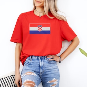 A woman wearing a red t-shirt with the Croatia national flag printed on it, standing against a white wall.