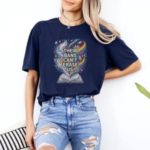 A woman wearing a navy blue t-shirt with a colorful graphic design and ripped jeans stands against a white wall, with a potted plant visible in the background.