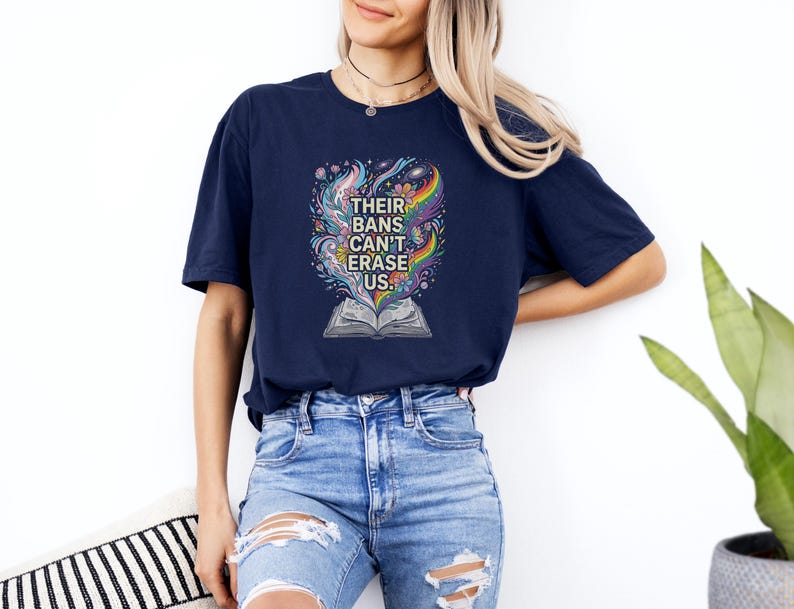 A woman wearing a navy blue t-shirt with a colorful graphic design and ripped jeans stands against a white wall, with a potted plant visible in the background.