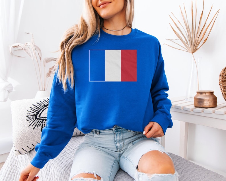 A woman wearing a blue sweatshirt with a French flag design is sitting on a bed in a room with a white wall and decorative elements.