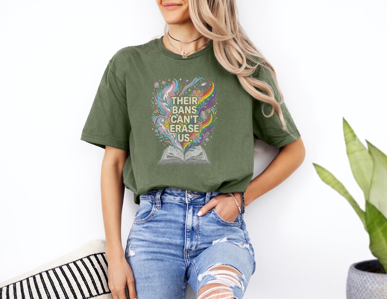 A woman wearing a green t-shirt with a colorful graphic and text, standing in front of a white wall with a potted plant to her right.