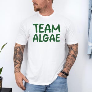 May include: A white short-sleeved t-shirt featuring the text "TEAM ALGAE" printed in a bold, dark green font across the chest. The shirt has a crew neckline and a standard fit, providing a clean and simple look for casual wear.