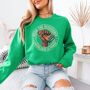 A woman wearing a green sweatshirt with a graphic design on it, sitting on a couch.