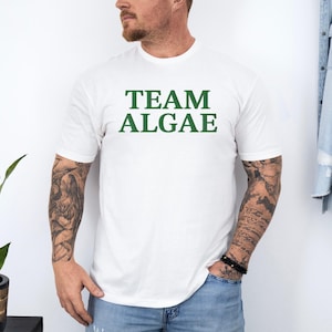 May include: A white short-sleeved t-shirt featuring the text TEAM ALGAE printed in dark green capital letters across the chest. The shirt has a crew neckline and a standard fit, providing a clean and simple look for casual wear.
