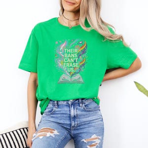 A woman wearing a green t-shirt with a colorful graphic and distressed jeans stands against a white wall, with a potted plant in the background.