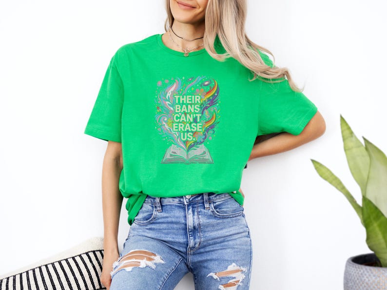 A woman wearing a green t-shirt with a colorful graphic and distressed jeans stands against a white wall, with a potted plant in the background.