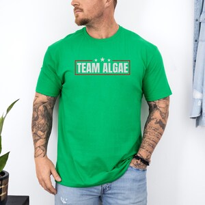 May include: A bright green short-sleeve t-shirt featuring a rectangular graphic on the chest. The graphic contains the text "TEAM ALGAE" in white capital letters, framed by a thin dark red border with three small white stars centered above the text.