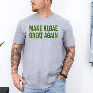 May include: A heather gray short-sleeved t-shirt featuring the text "MAKE ALGAE GREAT AGAIN" printed in a green, dripping, slime-like font across the chest. The shirt has a crew neckline and a standard fit, made of a soft, lightweight fabric.