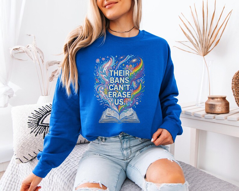 A woman wearing a blue sweatshirt with a colorful graphic design on it, sitting on a bed in a room with various decorative elements.