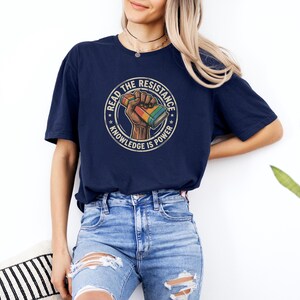 A woman wearing a navy blue t-shirt with a graphic design and jeans stands against a white wall, with a potted plant visible in the background.