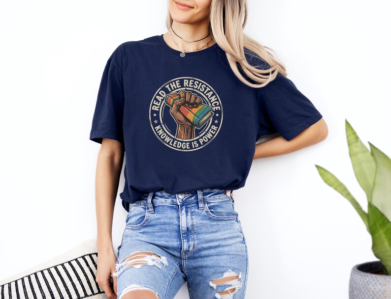 A woman wearing a navy blue t-shirt with a graphic design and jeans stands against a white wall, with a potted plant visible in the background.
