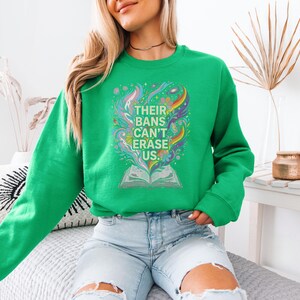 A woman wearing a green sweatshirt with a colorful graphic design and text printed on it, sitting on a couch in a room with decorative elements in the background.