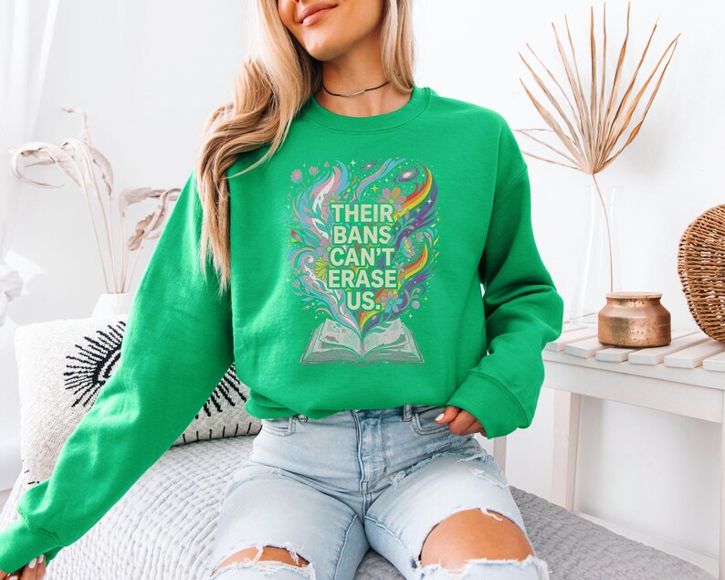 A woman wearing a green sweatshirt with a colorful graphic design and text printed on it, sitting on a couch in a room with decorative elements in the background.