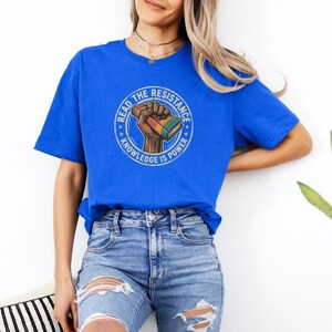 A woman wearing a blue t-shirt with a graphic design and jeans stands against a white wall, with a potted plant visible to her right.