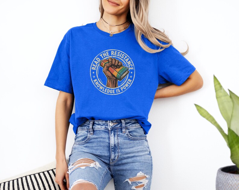 A woman wearing a blue t-shirt with a graphic design and jeans stands against a white wall, with a potted plant visible to her right.