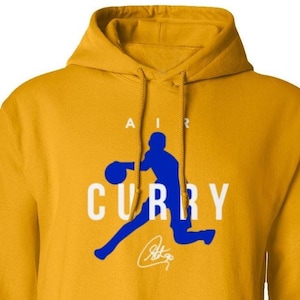May include: A yellow hoodie with a blue silhouette of a basketball player dribbling a ball. The text "AIR CURRY" is printed in white on the hoodie. The player's signature is printed below the text.
