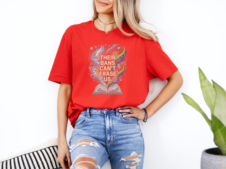 A woman wearing a red t-shirt with a colorful graphic and text, standing in front of a white wall with a potted plant to her right.