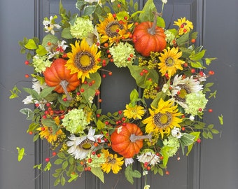 May include: A vibrant autumn wreath featuring sunflowers, orange pumpkins, and green foliage. The wreath is adorned with yellow and white flowers, red berries, and green hydrangeas. This decorative wreath is perfect for fall and seasonal home decor.
